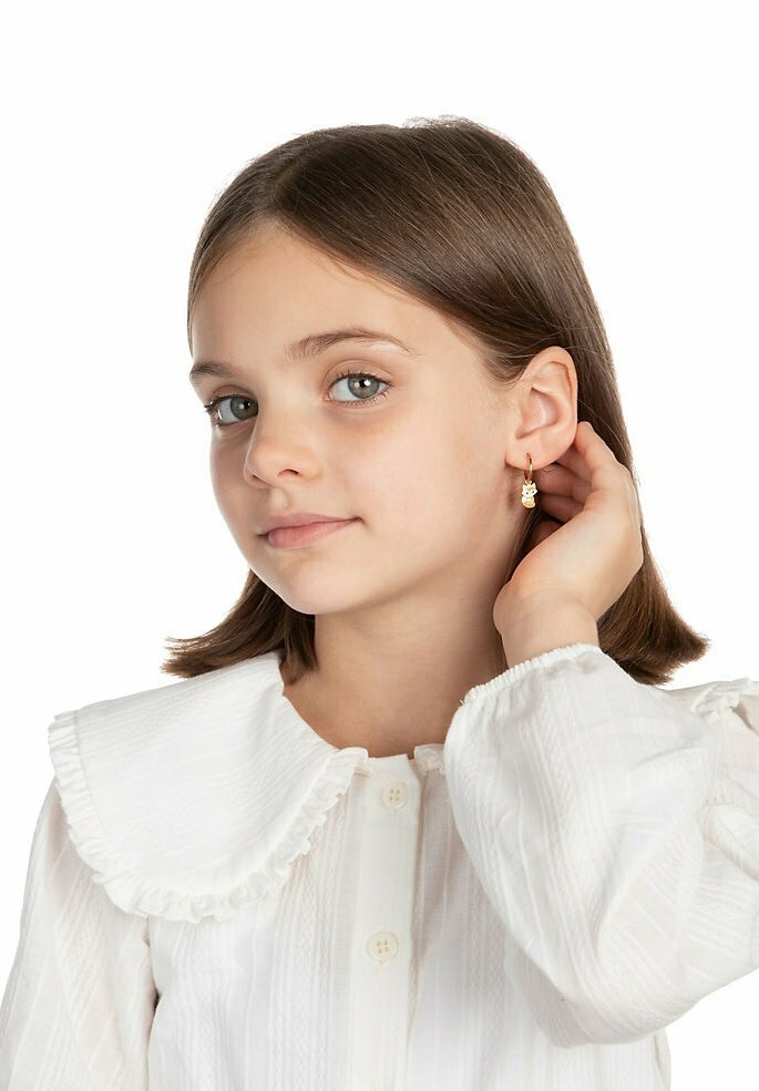 Goldene Creolen mit einem Anhänger, getragen von einem Mädchen. Sie hat glattes, braunes Haar und trägt eine weiße Bluse mit Kragen aus strukturiertem Stoff.