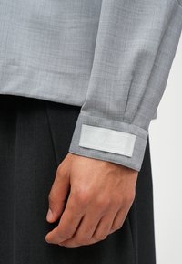 Chemise à manches longues grise avec une texture lisse, dotée d'une étiquette contrastante sur le poignet avec un texte brodé blanc.