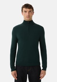 Mann trägt einen dunkelgrünen Pullover mit langen Ärmeln und halbem Reißverschluss sowie dunkle Hosen und steht vor einem schlichten weißen Hintergrund.