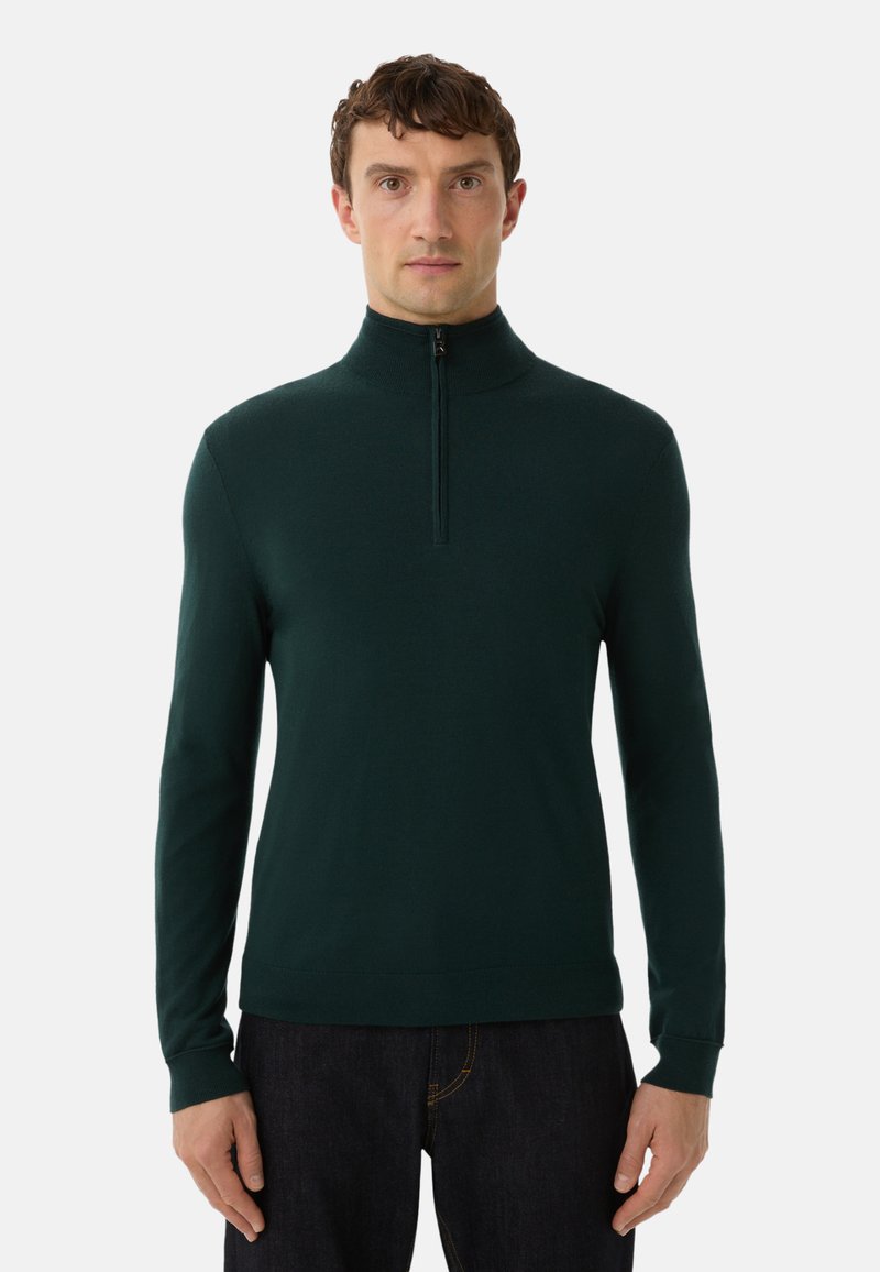 Mann trägt einen dunkelgrünen Pullover mit langen Ärmeln und halbem Reißverschluss sowie dunkle Hosen und steht vor einem schlichten weißen Hintergrund.