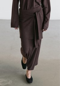 Person walking on concrete floor wearing a long, dark brown knit skirt and sweater with a tied belt and black flat shoes.