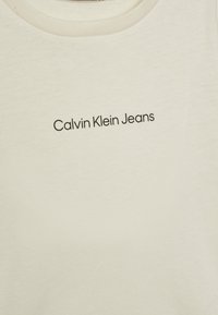 Krémový bavlněný tričko s klasickým kulatým výstřihem. Na středové části má černě tištěné logo "Calvin Klein Jeans". Hladký povrch.