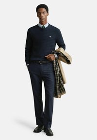 Námořnický pletený svetr přes košilovou košili, spárovaný se šedými kalhotami. Béžový kabát přehozený přes ruku, černé boty a hodinky.