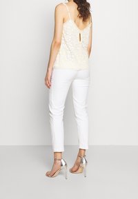 Haut cami en dentelle de couleur jaune pâle avec broderie florale, détail à l'arrière en forme de trou de serrure. Associé à un pantalon blanc slim et à des talons hauts argentés à lanières.