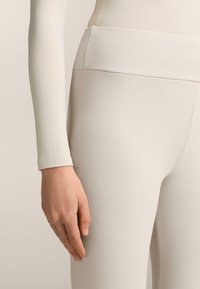 Lichtbeige leggings van rekbare stof, met een hoge tailleband en een gladde textuur. Hand rustend op het dijgebied.