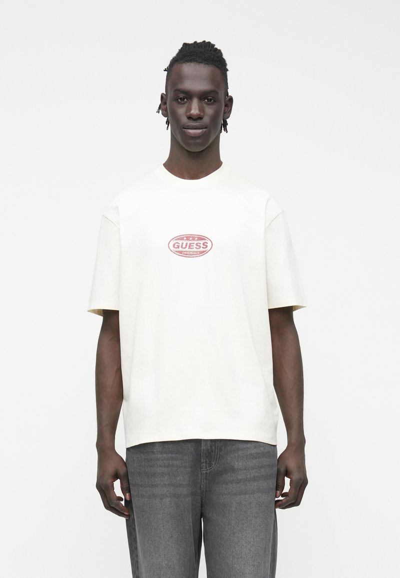 Camiseta de algodón color crema con cuello redondo y mangas cortas. Presenta un logo rojo y blanco "GUESS ORIGINALS" en el pecho. Combinada con vaqueros grises.