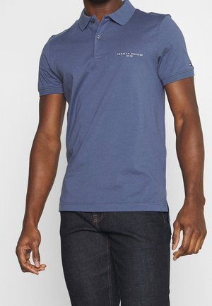 Man draagt een blauw Tommy Hilfiger poloshirt met logo op de borst en donkere spijkerbroek tegen een effen achtergrond.