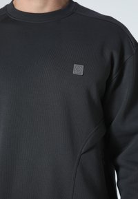 Felpa grigio scuro con scollo rotondo, dettagli testurizzati sulle spalle e una piccola toppa con il logo sul petto. Tessuto dalla texture liscia.