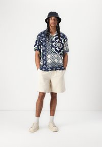Kurzärmeliges, navyfarbenes Hemd mit floralem Muster, kombiniert mit hellbeigen Shorts. Das Modell trägt einen schwarzen Bucket Hat und helle Sneaker.