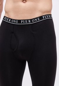 Torso masculin portant un pantalon de sous-vêtements longs noirs avec une ceinture marquée "PIER ONE" sur fond blanc.