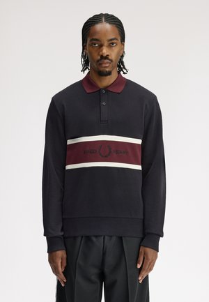 Mann mit geflochtenem Haar, der ein schwarzes Langarm-Poloshirt mit bordeauxrotem Kragen und Logo-Streifen über der Brust trägt, vor einem weißen Hintergrund steht.