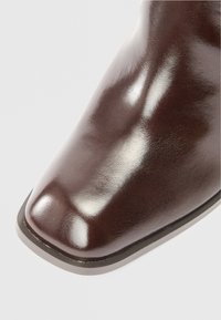 Close-up da bota de couro castanho-escuro brilhante, com o bico quadrado arredondado e a borda da sola contra um fundo claro.