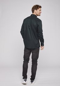 Dunkelgrünes langärmliges Hemd mit einem knöpfbaren Kragen, aus glattem Stoff. Getragen mit schwarzen Jeans und weißen Sneakern. Sichtbare Nähte.