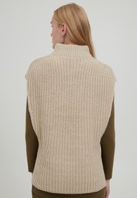 Beige gestrickte ärmellose Sweater mit hohem Kragen, ausgestattet mit einem strukturierten Muster und einer taillierten Silhouette, getragen über einem braunen Langarmtop.