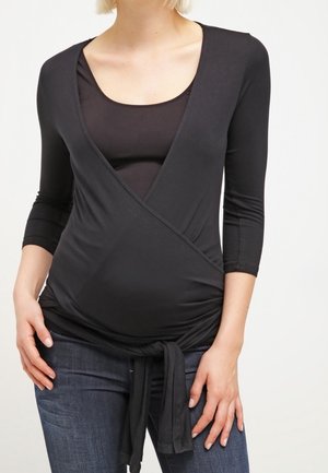 Mujer con una blusa premamá negra de estilo cruzado con mangas tres cuartos y vaqueros azul oscuro, con las manos apoyadas cerca de las caderas.