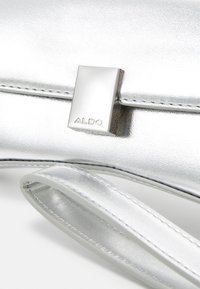 Stříbrná kovová kabelka s hladkou texturou, vybavená obdélníkovou stříbrnou sponou a odnímatelným popruhem, s vyraženým nápisem "ALDO."