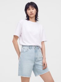 Jeune personne aux cheveux noirs portant un t-shirt blanc uni et un short en jean bleu clair sur un fond uni.
