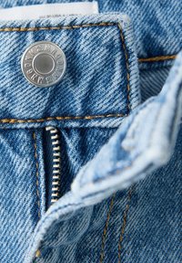 Gros plan d’un jean en denim bleu montrant un bouton métallique, les dents visibles de la fermeture éclair et des détails de couture jaunes.