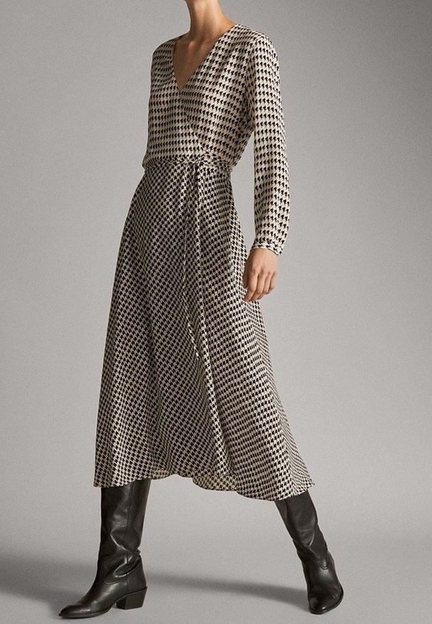 Femme portant une robe portefeuille à manches longues avec un motif pied-de-poule noir et blanc, associée à des bottes en cuir noir montant jusqu'aux genoux, debout devant un fond uni.