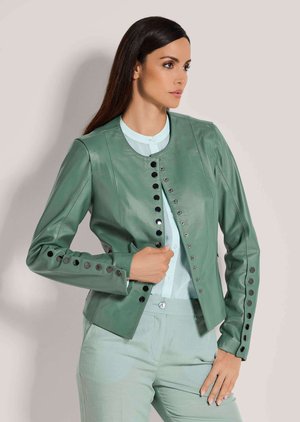 Frau mit langen dunklen Haaren, die eine hellgrüne Lederjacke mit schwarzen Knopfdetails und passende Hose trägt, posiert vor einem schlichten Hintergrund.