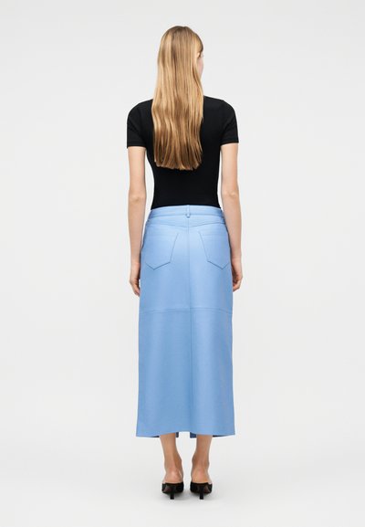Γυναίκα με μακριά ξανθά μαλλιά που φοράει μαύρο κοντομάνικο τοπ, ανοιχτό μπλε μίντι φούστα με πίσω τσέπες και μαύρα ψηλοτάκουνα παπούτσια, όρθια με την πλάτη προς τα εμπρός.