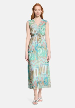 Jeune femme debout portant une robe portefeuille géométrique pastel sans manches avec un nœud à la taille et des sandales blanches sur un fond uni.