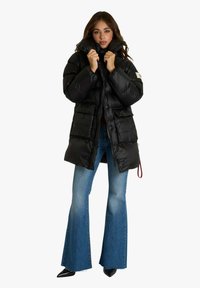 Giacca imbottita nera con collo alto, caratterizzata da una texture lucida, due tasche frontali e orlo con coulisse. Abbinata a jeans a zampa d'elefante.