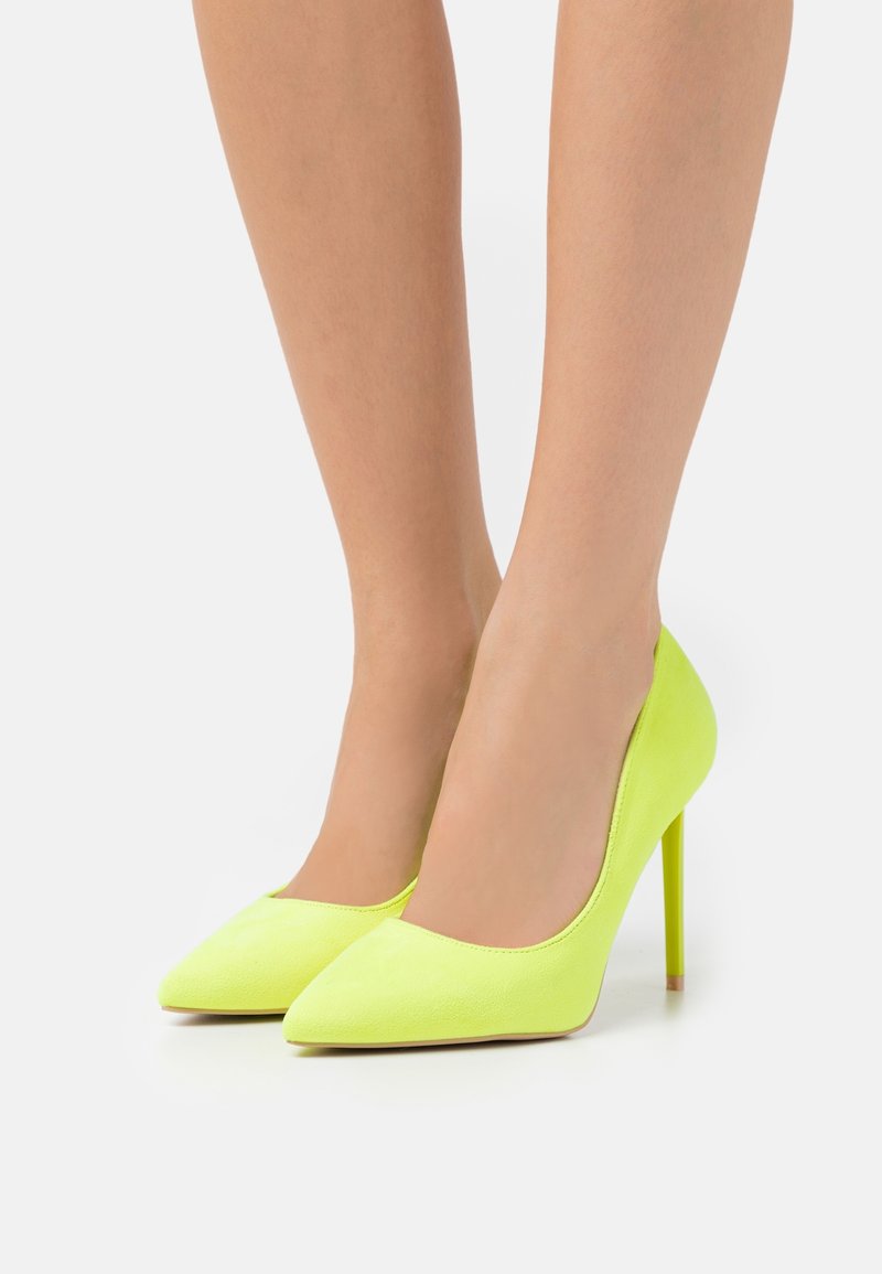 Jambes portant des escarpins à talons aiguilles pointus de couleur jaune vif sur un fond neutre.