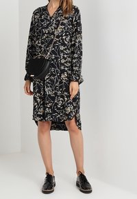 Musta kukallinen mekko pitkillä hihoilla, V-pääntiellä ja korkean-alhaisen helman muotoilulla. Yhdistetty mustien nauhallisten kenkien ja pienen pyöreän olkalaukun kanssa.