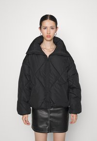 Chaqueta de plumón negra con un alto cuello, con cierre delantero, patrón de acolchado diagonal y mangas de gran tamaño. Usada sobre una falda de cuero negra.