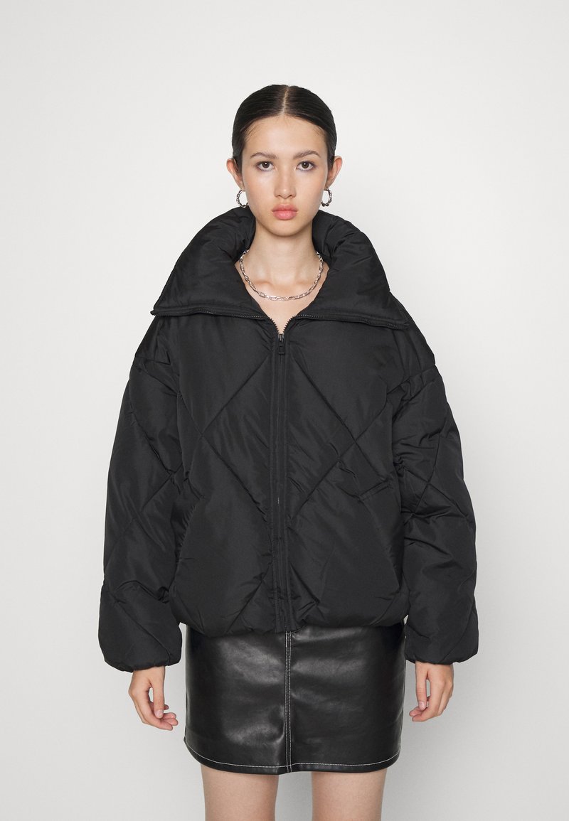 Chaqueta de plumón negra con un alto cuello, con cierre delantero, patrón de acolchado diagonal y mangas de gran tamaño. Usada sobre una falda de cuero negra.