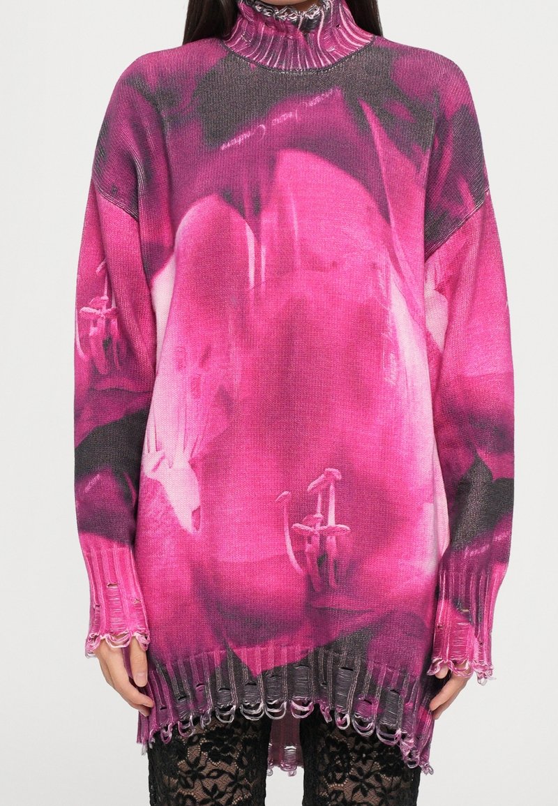 Femme portant un pull à col roulé côtelé long rose et noir avec ourlet et poignets usés, par-dessus un legging en dentelle noire.