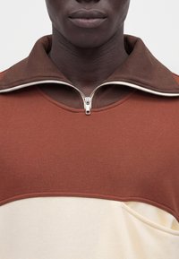 Gros plan d'une personne portant un haut marron et beige zippé avec la fermeture partiellement ouverte au niveau du col.