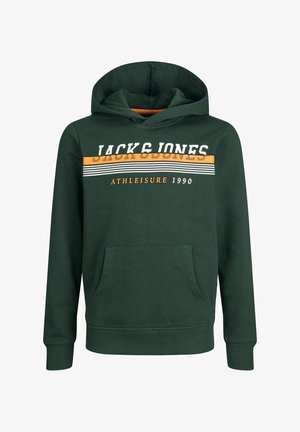 Osoba ubrana w ciemnozieloną bluzę z kapturem z napisem "Jack & Jones Athleisure 1990", niebieskie dżinsy i granatowo-białe trampki, stoi z rękami w kieszeniach.