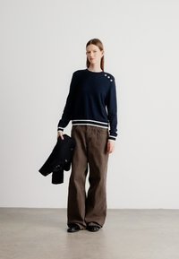 Μπλε πουλόβερ πλεκτό με ρίγα στο τελείωμα, με τρία κουμπιά στον ώμο. Συνδυάζεται με φαρδιά καφέ παντελόνια. Το μοντέλο κρατά ένα μπλε σακάκι.