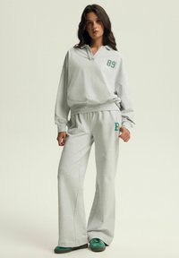 Jasnoszara, oversize'owa bluza z zielonym nadrukiem "89!" w komplecie ze spodniami dresowymi w tym samym kolorze z zielonym "B." Płaskie buty w kolorze teal.