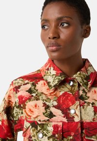 Kurtka w kwiatowy wzór z czerwonym, różowym i zielonym motywem, z kołnierzem, zatrzaskami i naszywanymi kieszeniami z przodu.