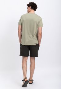 Lichte olijfgroene T-shirt met korte mouwen, gecombineerd met donkere shorts. Model staat met de rug naar de camera, met bruine sandalen aan.