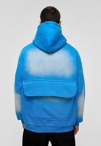 Overmaatse blauwe hoodie met een verloop van felblauw naar lichtblauw, voorzien van een grote achterzak en structuurtstof.
