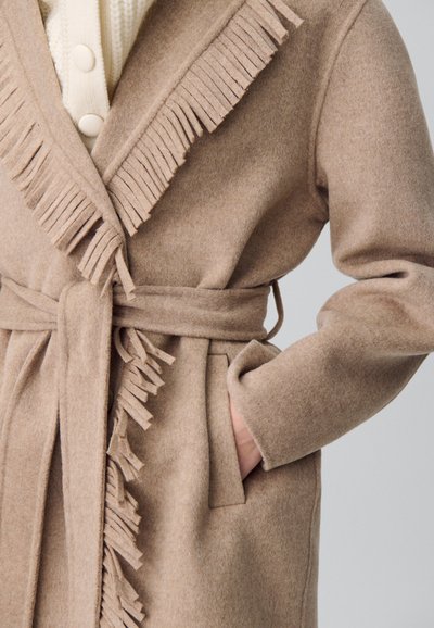 Manteau en laine beige avec une ceinture et des franges le long du col et des bords avant, porté sur un pull crème à boutons.