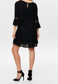 Vestido negro con mangas tres cuartos, puños con volantes, detalles de encaje y un aro en la parte trasera. Combinado con zapatos de tacón alto negros.