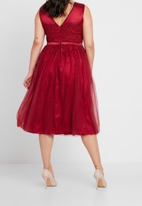 Rode tule jurk met een getailleerde beha met kantdetails en een V-rug, vloeiende rok en een satijnen tailleband. Inclusief een paar beige hakken.