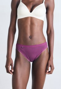 Femme avec la peau foncée portant un soutien-gorge blanc et des sous-vêtements violets, se tenant contre un fond clair uni.
