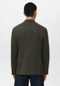 Temnozeleni herringbone blazer z odrezanimi reverji, dvema zadnjima odprtinama in enojnimi gumbnimi manšetami, viden s hrbta. Gladka tekstura.