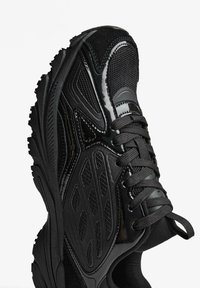 Crna atletska cipela s teksturiranim gornjim dijelom, glatkim lakiranim detaljima, zatvaranjem na vezice i masivnim gumennim potplatom s izraženim uzorkom.