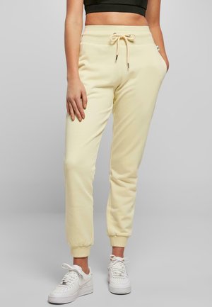 Femme portant un pantalon de jogging crème avec une taille à cordon de serrage et des baskets blanches, debout devant un fond gris uni.