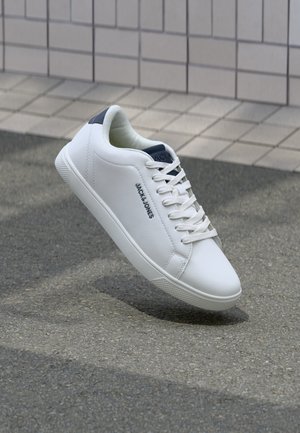 Adidași din piele albă cu un design low-top, suprafață texturată, accent la călcâi de culoare albastră închis și o talpă plată din cauciuc cu un model de profil.