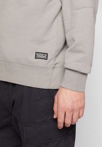 Sudadera gris claro con un acabado texturizado. Presenta una etiqueta negra de marca en el dobladillo. Se lleva con pantalones cargo oscuros.