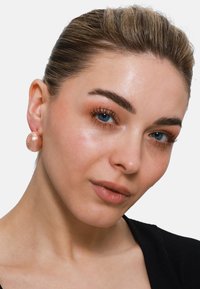 Orecchini a bottone rotondi in oro rosa, con una superficie liscia, indossati da un modello, caratterizzati da un sottile motivo. Il design è audace e contemporaneo.