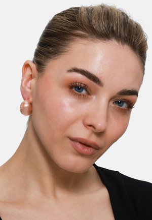 Orecchini a bottone rotondi in oro rosa, con una superficie liscia, indossati da un modello, caratterizzati da un sottile motivo. Il design è audace e contemporaneo.